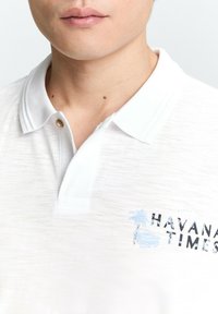 Weißes Poloshirt mit strukturiertem Finish, das ein blaues Palmenbaum-Motiv und schwarzen "HAVANA TIMES"-Text aufweist. Der Kragen hat einen einzelnen goldenen Knopf.