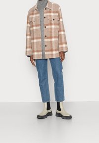 Persona vistiendo una chaqueta de cuadros en beige y blanco, un suéter de cuello alto gris, jeans azules y botas de tobillo beige con lados elásticos negros.