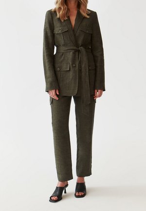 Nainen, joka pukeutunut oliivinvihreään, vyölliseen bleiseriin, jossa on läpätaskut, ja yhteensopiviin suoralahkeisiin housuihin, yhdistettynä mustiin avoimella varpaalla varustettuihin korkokenkiin.