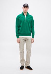 Zielony pulower z półgolfem na zamek, długimi rękawami i ściągaczami, zestawiony z jasnobeżowymi jeansami oraz czarnymi sneakersami.