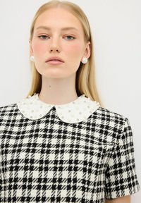 Schwarz-weiß karierte Kleid mit einem Kragen, der mit weißen Perlen akzentuiert ist. Der Stoff hat eine strukturierte Oberfläche und das Design hat kurze Ärmel.