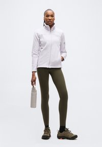 Γυναίκα στέκεται κρατώντας ένα γκρι παγούρι, φοράει ένα ανοιχτό μωβ φλις με φερμουάρ, λάδι κολάν, μαύρες κάλτσες και παπούτσια πεζοπορίας.