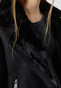 Černá umělá kožešinová bunda s měkkou texturou, se zipem vpředu a širokým límcem. Detailní záběr na látku a detaily.