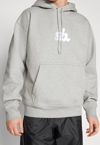 Hombre con sudadera ligera gris con el logo blanco "sb." y pantalones casuales negros, de pie con las manos relajadas a los lados frente a un fondo sencillo.