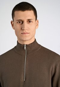 Brauner Zip-Jumper mit hohem Kragen und strukturiertem Strick, ausgestattet mit einem silbernen Reißverschluss und einer taillierten Passform. Glattes Material mit minimaler Gestaltung.
