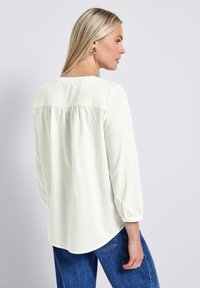 Blusa bianca testurizzata con scollatura a barchetta, maniche a tre quarti e orlo curvo, abbinata a jeans in denim blu scuro.