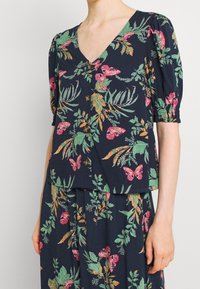 Donna che indossa un top e una gonna blu navy con motivi floreali di farfalle rosa e foglie verdi, dotati di maniche corte e bottoni frontali.