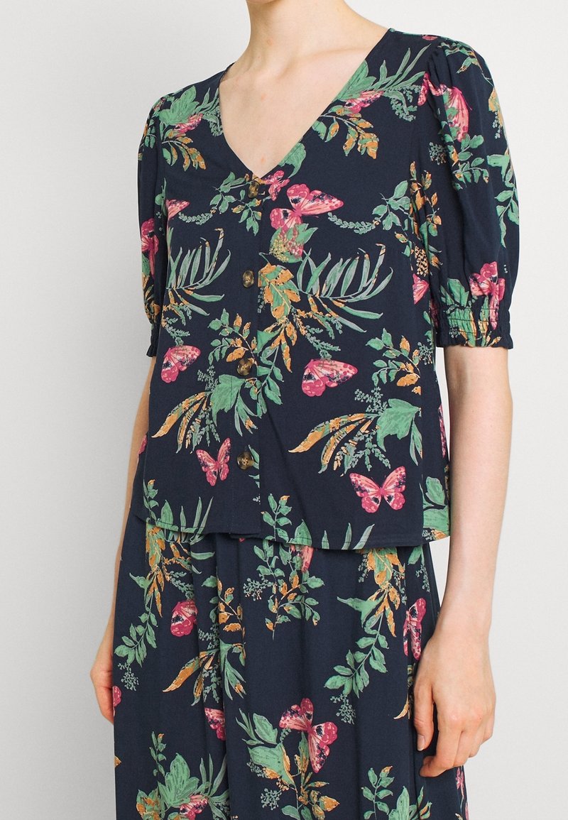 Donna che indossa un top e una gonna blu navy con motivi floreali di farfalle rosa e foglie verdi, dotati di maniche corte e bottoni frontali.