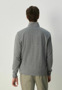 Pulover gri, în dungi, cu guler înalt și mâneci lungi. Textură moale, model cordat, design ajustat, purtat cu pantaloni de culori deschise.