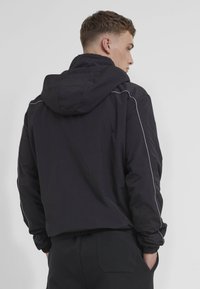 Black hooded jacket featuring elastic cuffs and drawstring hem. Linear silver accents along the sleeves add detail. Soft, textured fabric.