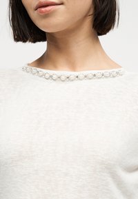 Γυναίκα που φοράει λευκό πλεκτό τοπ με στρογγυλή λαιμόκοψη διακοσμημένη με σειρά από μεγάλες πέρλες και μικρές ασημένιες λεπτομέρειες.