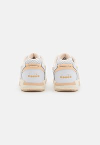 White and tan sneakers with a textured fabric back, circular perforations, and a gold logo at the heel. Rubber sole with a light design.