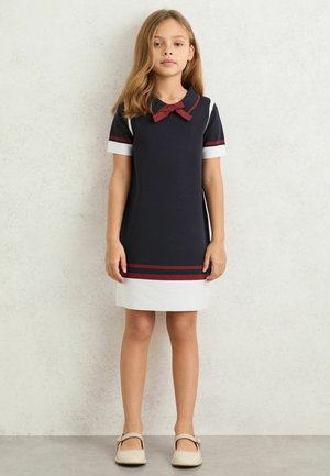 Rochie cu mâneci scurte în albastru închis și alb, cu guler rotund și fundă bordo. Prezintă dungi orizontale roșii la tiv și pe mâneci.