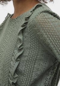 Top verde in pizzo con motivi intricati, tessuto testurizzato, dettaglio a volant lungo la spalla e un design a maglia aperta sulle maniche.