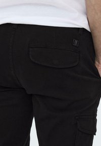 Pantalons cargo noirs avec une poche latérale et une poche arrière avec une étiquette en tissu. Le matériau semble robuste avec une texture lisse.