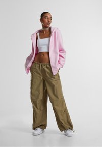 Svetloružová zipová mikina preložená cez biely crop top, spojená s voľnými olivovými cargo nohavicami a bielymi teniskami.