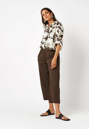 Woman wearing a white floral shirt, brown cropped pants, black sandals, and large earrings standing against a white background.