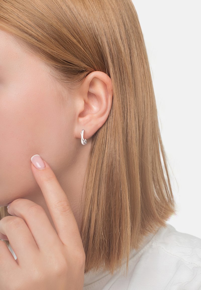 Silver vriden hoopearring med en polerad yta, som visar en slank design. Mjukt, naturligt hår och neutral nagellack synligt.