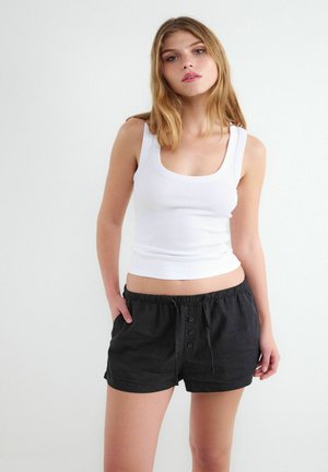 Jeune femme portant un haut blanc sans manches et un short noir ample, debout avec une main dans la poche devant un fond clair uni.