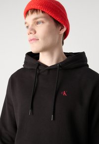 Melnā kapuce ar auklu, ar nelielu sarkanu "CK" logotipu. Modelis valkā teksturētu sarkanu adītu cepuri. Gluda kokvilnas auduma.