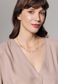 Złoty naszyjnik z podwójnym sznurem, ozdobiony kolorowymi koralikami w odcieniach różu, czerni i zieleni, noszony na miękkiej beżowej odzieży z dekoltem w serek.