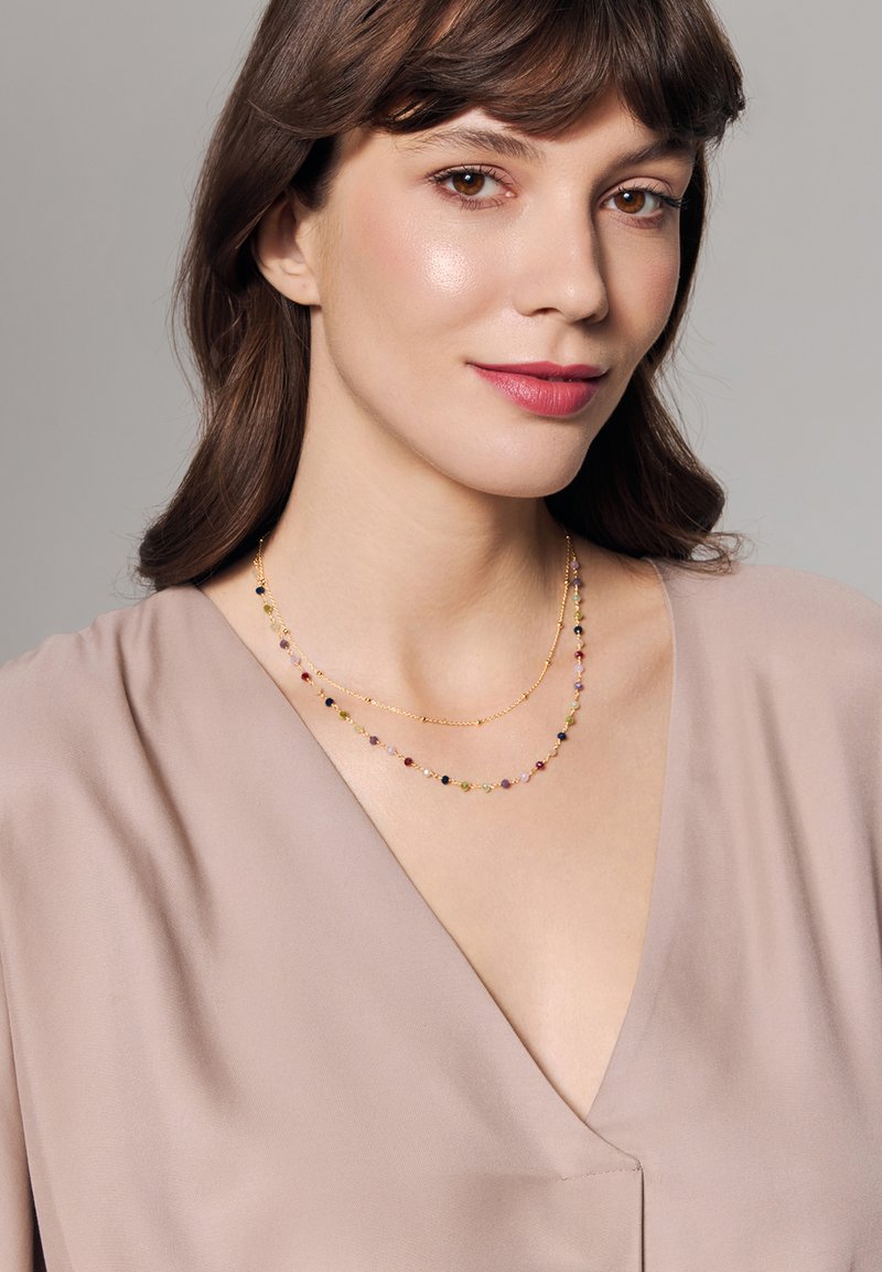 Złoty naszyjnik z podwójnym sznurem, ozdobiony kolorowymi koralikami w odcieniach różu, czerni i zieleni, noszony na miękkiej beżowej odzieży z dekoltem w serek.