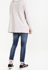 Ljusgrå långärmad hoodie med ett texturerat mönster, kombinerad med blå denimjeans. Jeansen är upprullade vid fållarna och bärs med svarta sneakers.