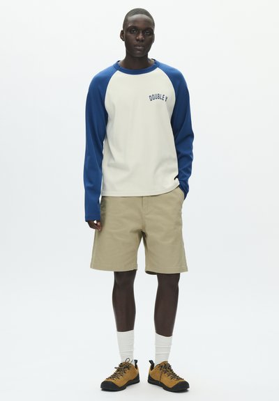 Chemise crème à manches longues avec manches raglan bleues et logo, associée à un short beige. Chaussures de randonnée noires et jaunes avec des chaussettes blanches.