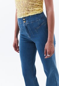 Vaqueros de mezclilla azul de cintura alta con un corte ajustado, que cuentan con dos botones frontales y un diseño estructurado sutil. Tejido texturizado con costuras mínimas.