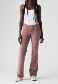 Jean évasé en velours côtelé rose avec une fermeture à bouton et zip, poches avant, et un tissu doux et texturé, assorti à un débardeur blanc et des baskets.