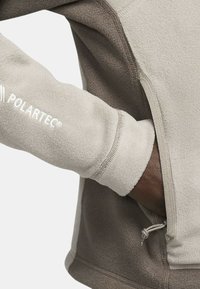 Giacca in pile con una combinazione di colori grigio-marrone, caratterizzata da una tasca con zip, fori per le dita e il logo Polartec sul braccio. Texture liscia.