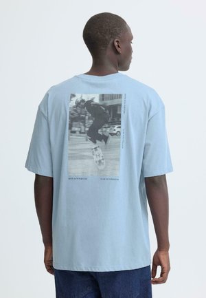 Hellblaues Baumwoll-T-Shirt mit einem Grafikdruck auf der Rückseite, der einen Skateboarder zeigt, mit markanten Textphrasen und einer lockeren Passform.