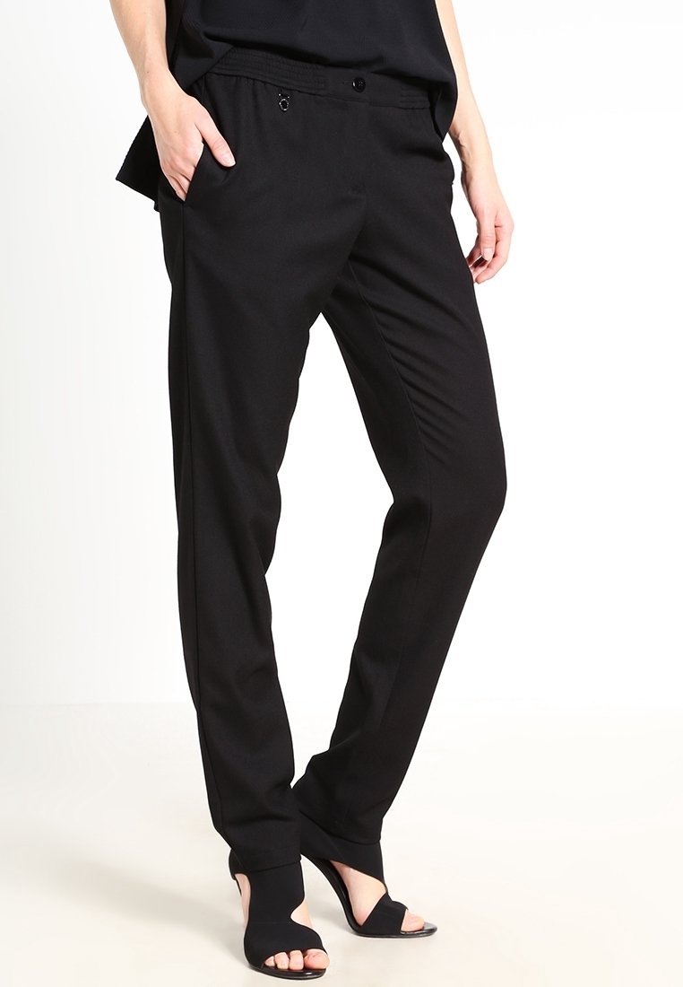 Femme portant un pantalon noir fuselé avec des poches et des chaussures à talons ouverts noires, debout avec une main dans la poche.