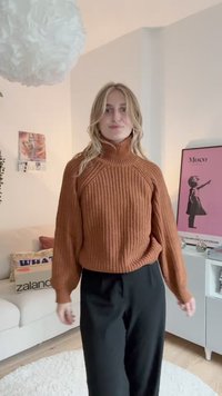Rust-orange Strickpullover mit hohem Kragen, strukturiertem Muster und lässiger Passform, kombiniert mit schwarzen, weit geschnittenen Hosen. Schlichter weißer Hintergrund.