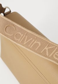 Beżowa torebka o wyraźnie zarysowanym kształcie. Charakteryzuje się fakturowanym wykończeniem oraz szerokim, haftowanym paskiem z napisem „Calvin Klein”.