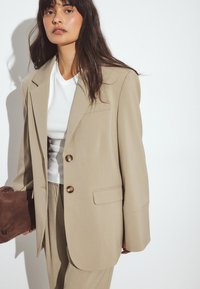 Beige oversized blazer med et struktureret design, to guldknapper og frontlommer. Parret med en hvid top og en brun clutch.