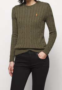 Olivgrön kabelstickad tröja med rund halsringning och ribbad fåll; har en liten orange logotyp på vänster bröst. Matchas med svarta jeans.