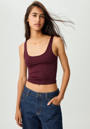 Ung kvinde med langt mørkt hår iført øreringe som hoops, en ærmeløs crop top i burgunder og blå jeans, stående med den ene hånd i lommen.