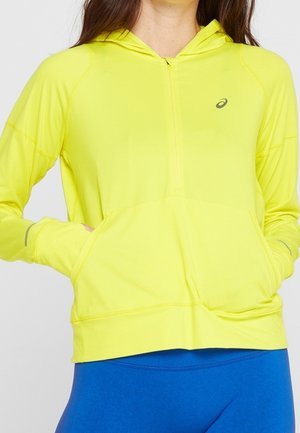 Person trägt ein hellgelbes Kapuzen-Sweatshirt mit Reißverschluss und Vordertaschen sowie blaue Sportleggings vor einfachem Hintergrund.