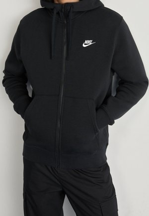 Personne portant un sweat à capuche noir Nike zippé, avec des cordons et des poches avant, assorti à un pantalon noir.