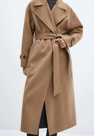 Kobieta ubrana w długi płaszcz w kolorze wielbłądzim z paskiem i szerokimi klapami, na ciemnej prążkowanej sukience, stojąca na jednolitym tle.