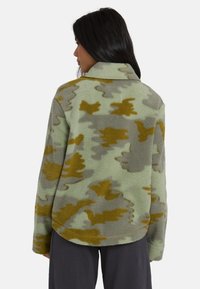 Donna con capelli lunghi e scuri che indossa una giacca in pile a scacchi verde, grigio e senape, vista da dietro, su uno sfondo neutro.