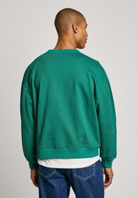 Sudadera verde de corte relajado, con cuello redondo y puños acanalados, superpuesta sobre una camisa blanca, combinada con jeans azules. Textura suave y lisa.