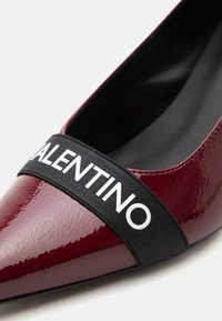 Chaussure pointue rouge foncé brillante avec une bride noire sur l'avant comportant l'inscription blanche "VALENTINO".