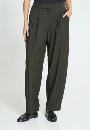 Vrouw draagt hoog getailleerde donkerolijfgroene wijde broek met plooien, zwarte loafers en één hand in de zak.