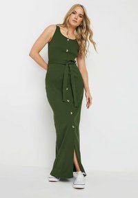 Robe maxi verte à rayures avec débardeur, détails de boutons, ceinture à la taille et fente sur le côté. Associée à des baskets blanches. Texture lisse.
