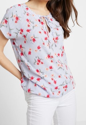 Femme portant un chemisier à manches courtes à rayures bleues et blanches avec un imprimé floral rouge, associé à un pantalon blanc sur un fond uni.