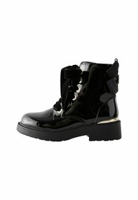 Botas de tobillo de charol negro con suela gruesa, ojales dorados y un moño decorativo en la parte trasera. Los cordones van acompañados de una cinta gruesa.
