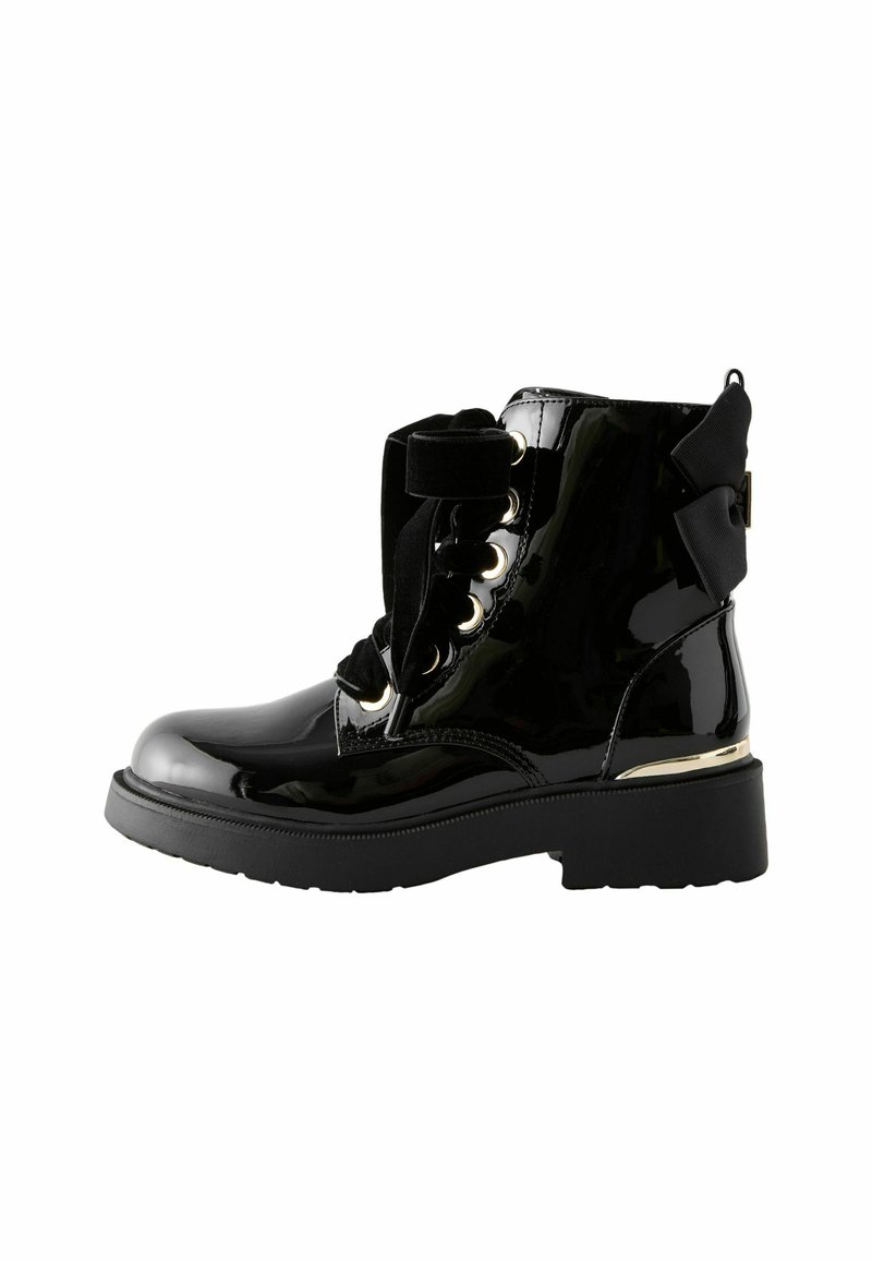 Botas de tobillo de charol negro con suela gruesa, ojales dorados y un moño decorativo en la parte trasera. Los cordones van acompañados de una cinta gruesa.