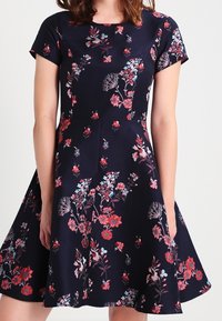 Šaty s květinovým potiskem a krátkými rukávy, s upnutým korzetem a rozšířenou sukní. Hlavní barva je námořnická, s červenými a modrými květinovými vzory.